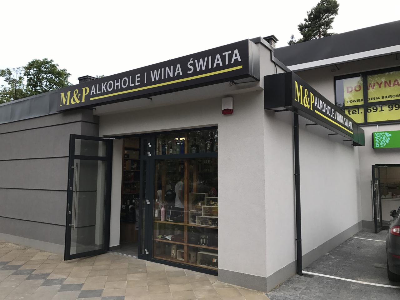 Fasada budynku z szyldem 'M&P ALKOHOLE I WINA ŚWIATA' nad otwartymi drzwiami wejściowymi, widoczne półki z butelkami wewnątrz.