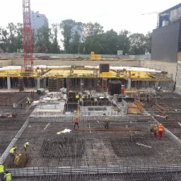 Rozległy plac budowy z widoczną konstrukcją zbrojeniową, żółtymi elementami szalunkowymi i pracującymi robotnikami w kamizelkach odblaskowych, na tle miejskiego krajobrazu z dźwigiem i budynkami.