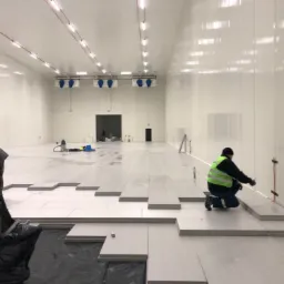 Montaż paneli podłogowych w chłodni, widoczni pracownicy w odzieży roboczej i elementy instalacji chłodniczej pod sufitem.