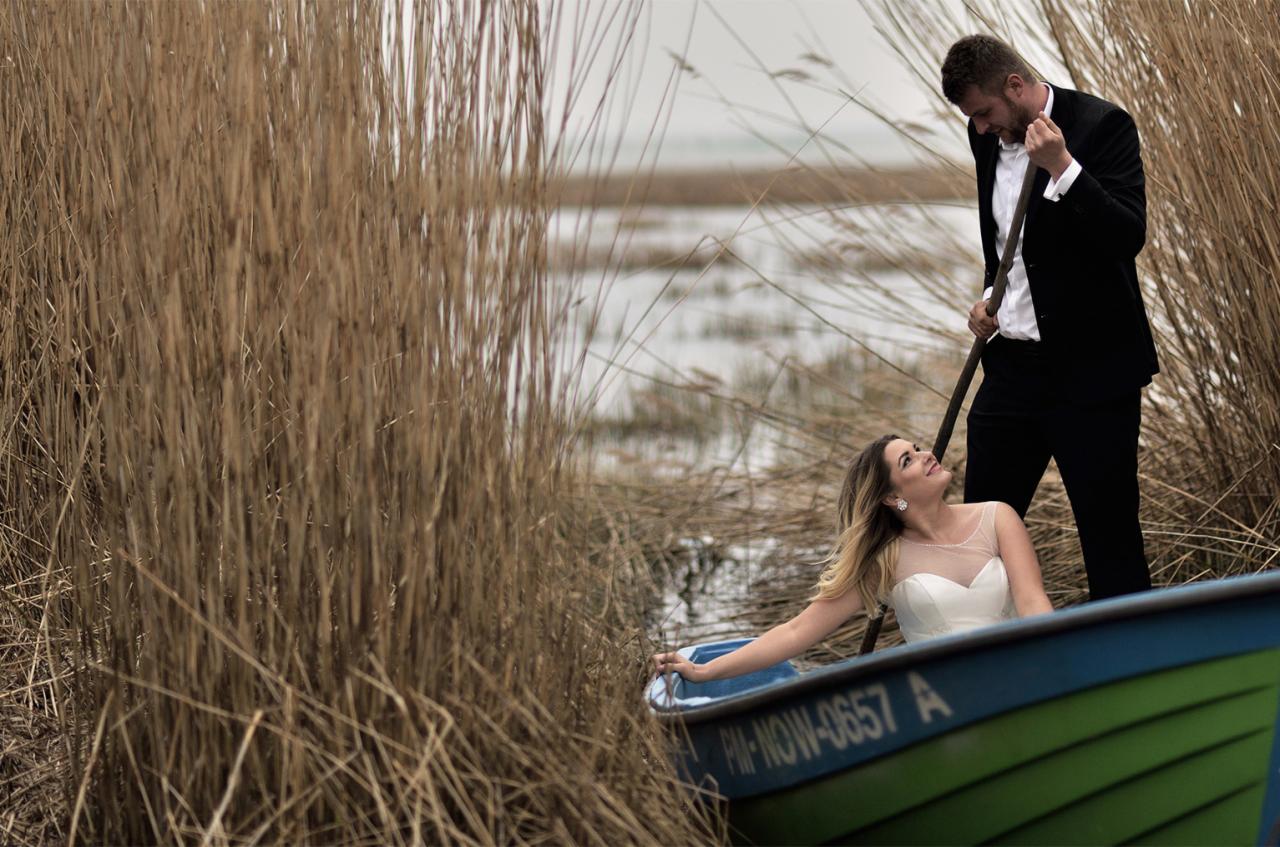 Para młoda w łódce na tle trzcin: panna młoda w białej sukni patrzy z uśmiechem na pana młodego w czarnym garniturze, który trzyma wiosło.