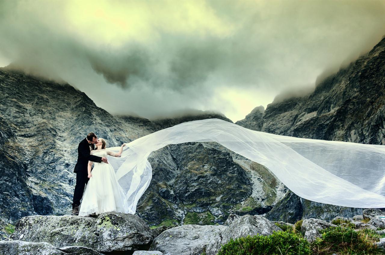 Para młoda całuje się na szczycie skalistego wzgórza z rozwianym welonem na tle górskiego krajobrazu i pochmurnego nieba.