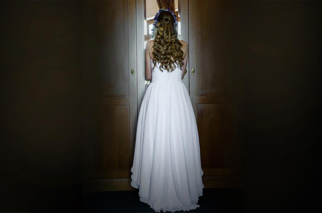 Panna młoda w białej sukni, widziana od tyłu, stoi przed uchylonymi, ciemnymi drzwiami, z wiankiem z fioletowych kwiatów we włosach.
