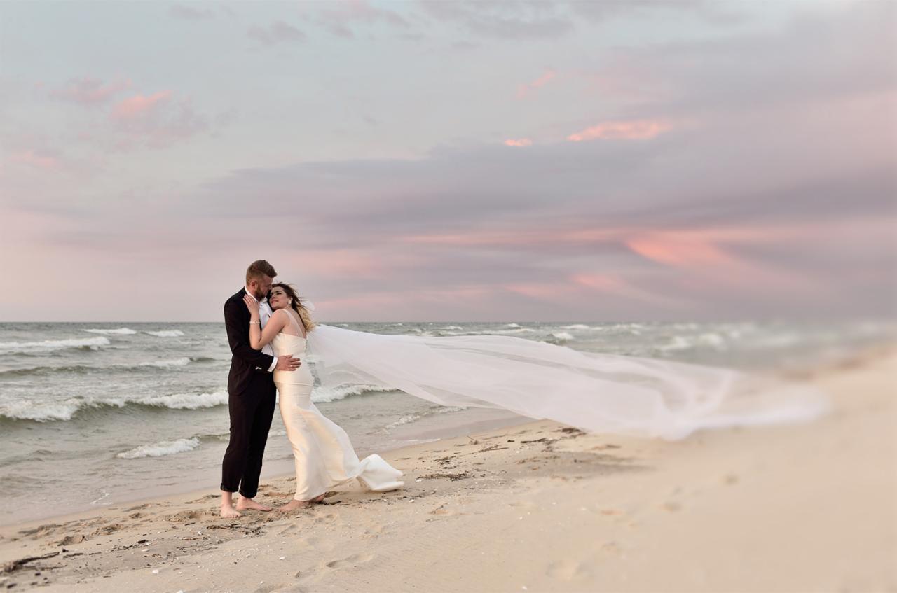 Para młoda na plaży o zachodzie słońca, panna młoda w dopasowanej sukni z długim welonem, pan młody w czarnym smokingu, oboje boso na piasku, fale morskie w tle.