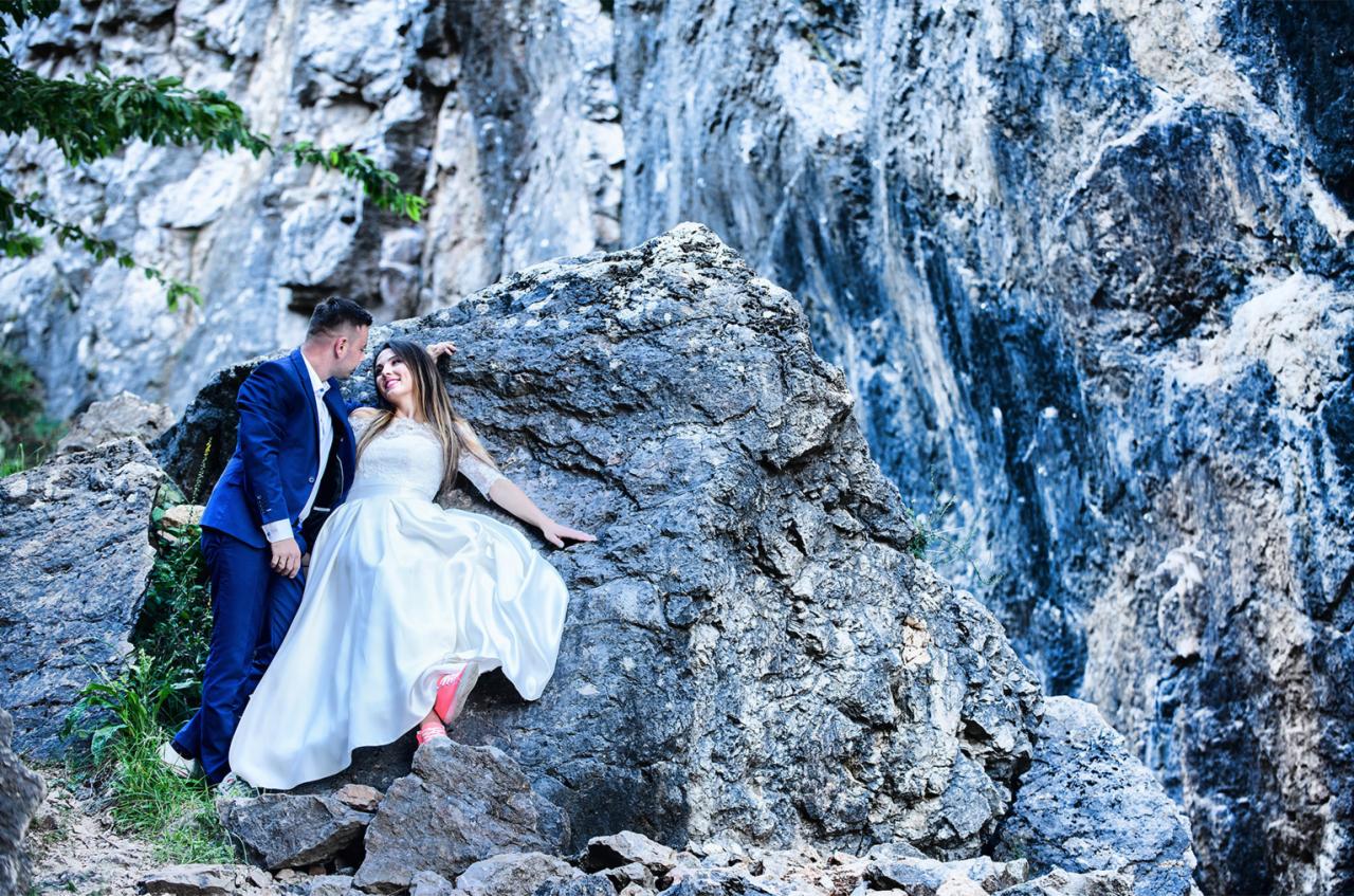 Para młoda w strojach ślubnych pozuje na tle skalnej ściany. Panna młoda w białej sukni z koronkową górą i satynowym dołem, opiera się o duży głaz. Pan młody w granatowym garniturze stoi obok...