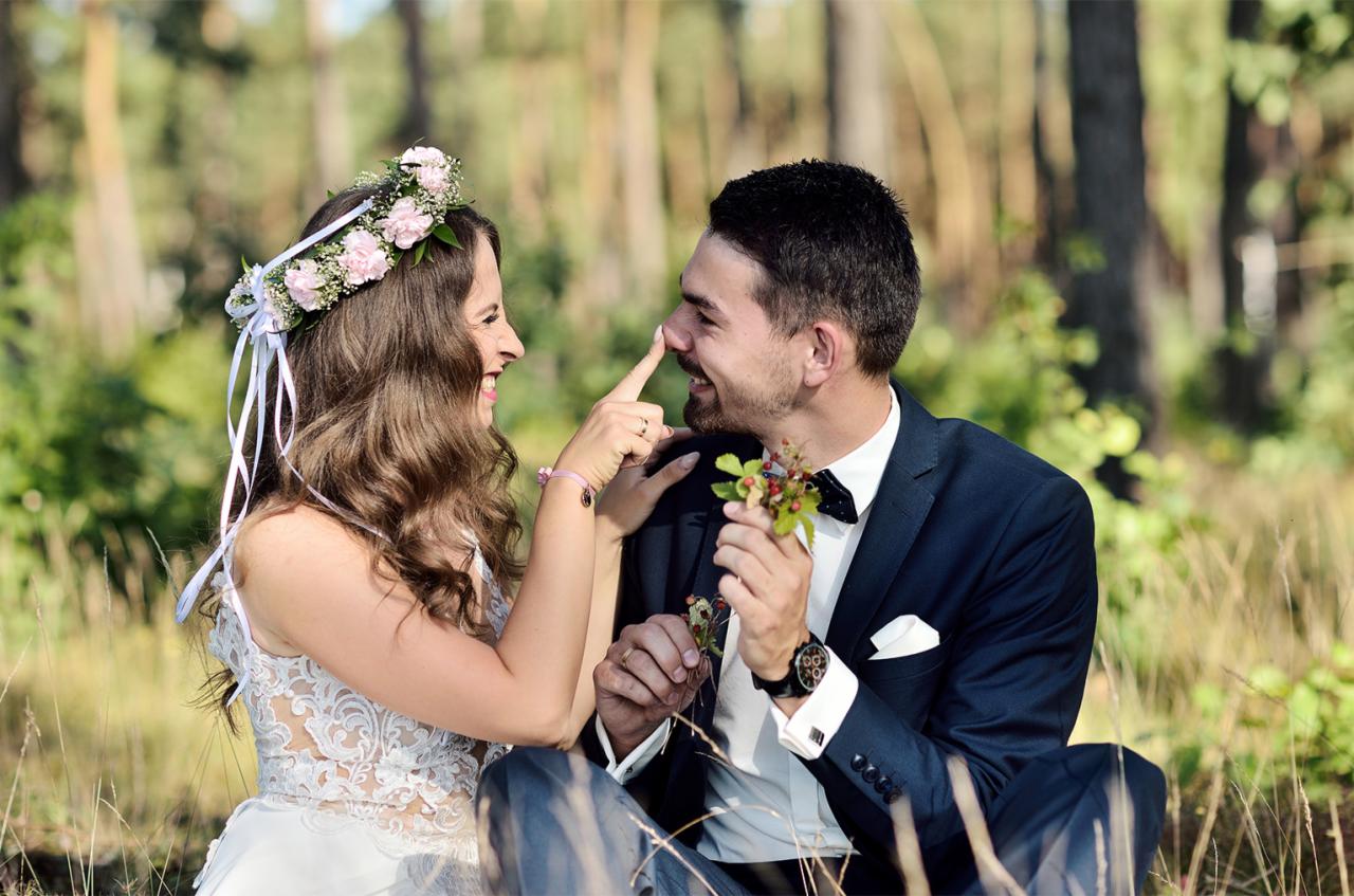 Panna młoda w wianku z róż dotyka palcem nosa uśmiechniętego pana młodego w granatowym garniturze, siedzących na tle leśnej zieleni.