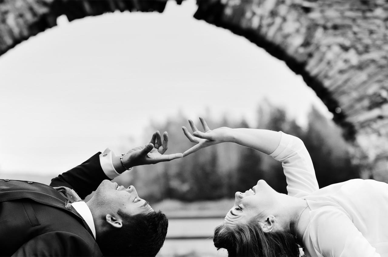 Czarno-białe zdjęcie pary leżącej na plecach, twarzami do siebie, z dłońmi ułożonymi w kształt serca na tle kamiennego łuku i drzew.