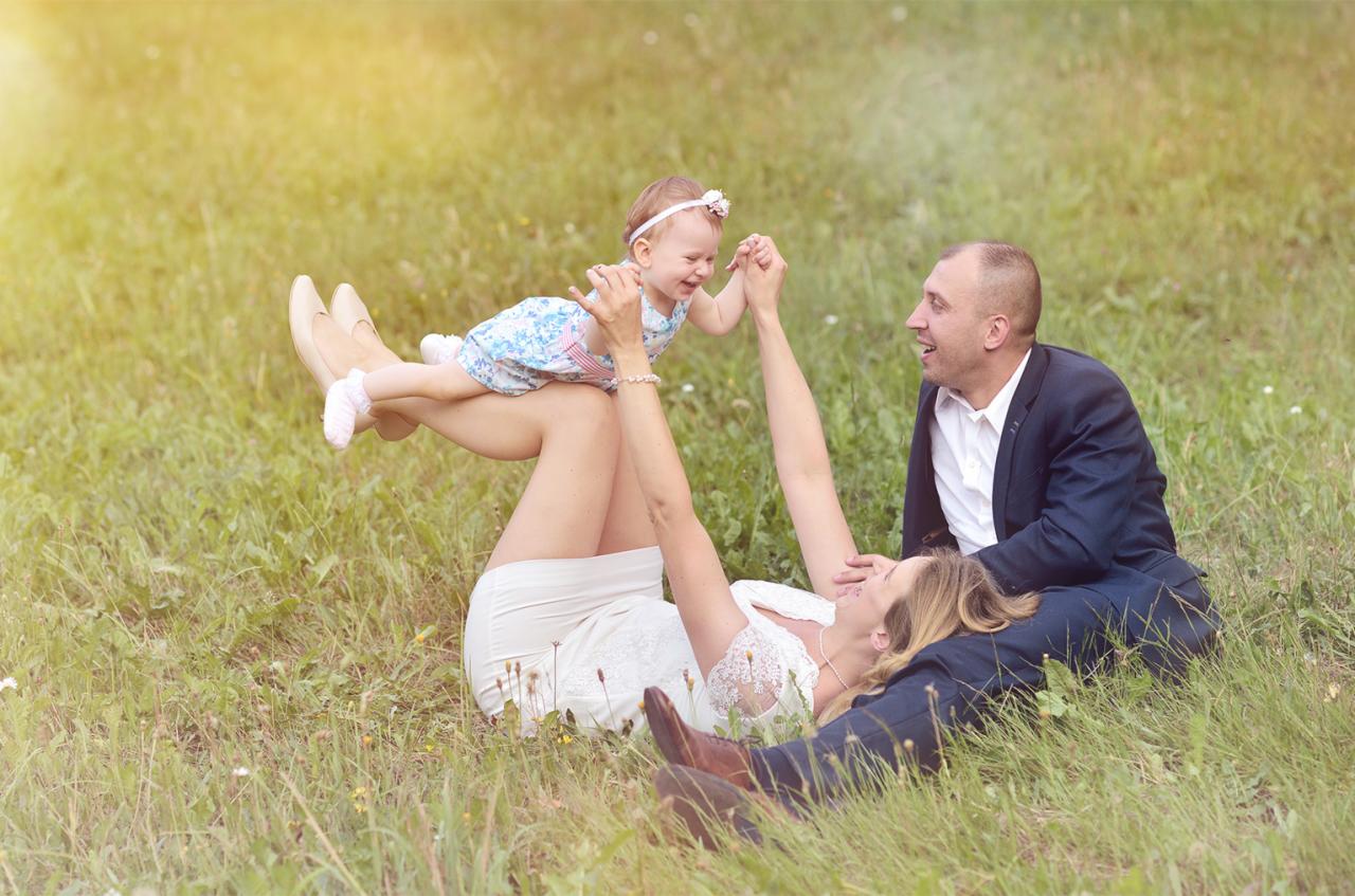 Szczęśliwa rodzina na łące: mama leży na trawie, trzymając uśmiechniętą córeczkę w górze, tata siedzi obok, patrząc z radością.