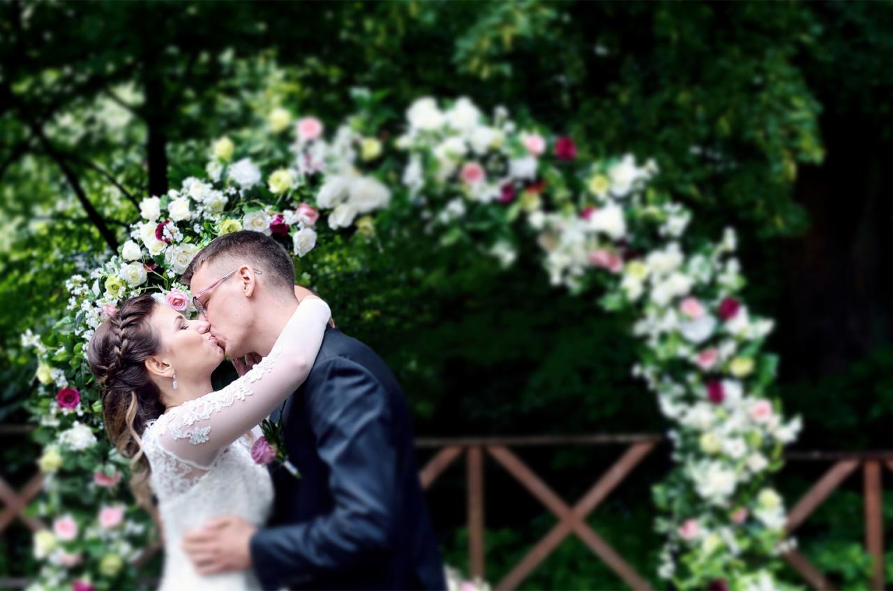 Pocałunek nowożeńców na tle kwiatowej bramy z białych i różowych róż, panna młoda w koronkowej sukni, pan młody w ciemnym garniturze i różowych okularach.