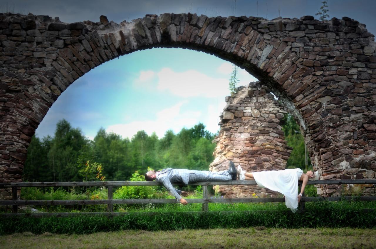 Para młoda w nietypowej pozie leży na drewnianym płocie na tle kamiennego łuku i zieleni, tworząc surrealistyczny obraz