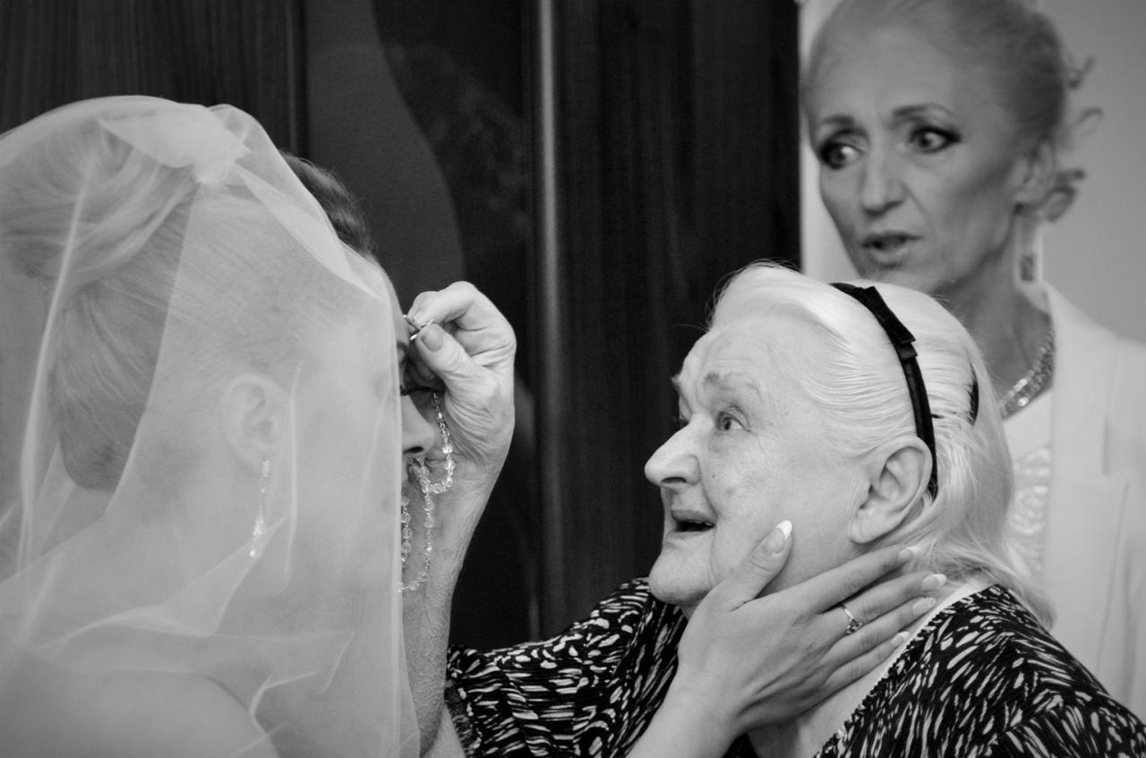 Czarno-białe zdjęcie przedstawia przygotowania panny młodej do ślubu: starsza kobieta w czarno-białej bluzce poprawia welon i kolczyki młodej kobiety, w tle widoczna inna kobieta z zaniepokojonym...