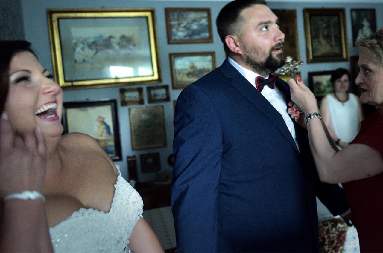 Przygotowania do ślubu: panna młoda śmieje się, podczas gdy inna kobieta poprawia muszkę panu młodemu w granatowym garniturze, w tle ściana z licznymi obrazami w złotych ramach.