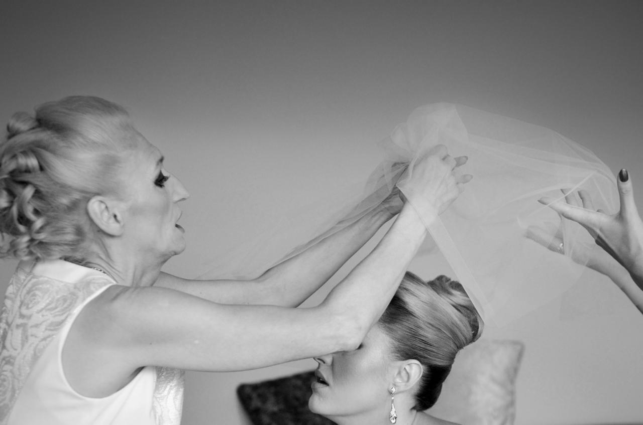Czarno-białe zdjęcie przedstawia matkę pomagającą pannie młodej założyć welon, widoczne dłonie trzymające delikatną tkaninę, elegancka fryzura panny młodej i wiszące kolczyki.