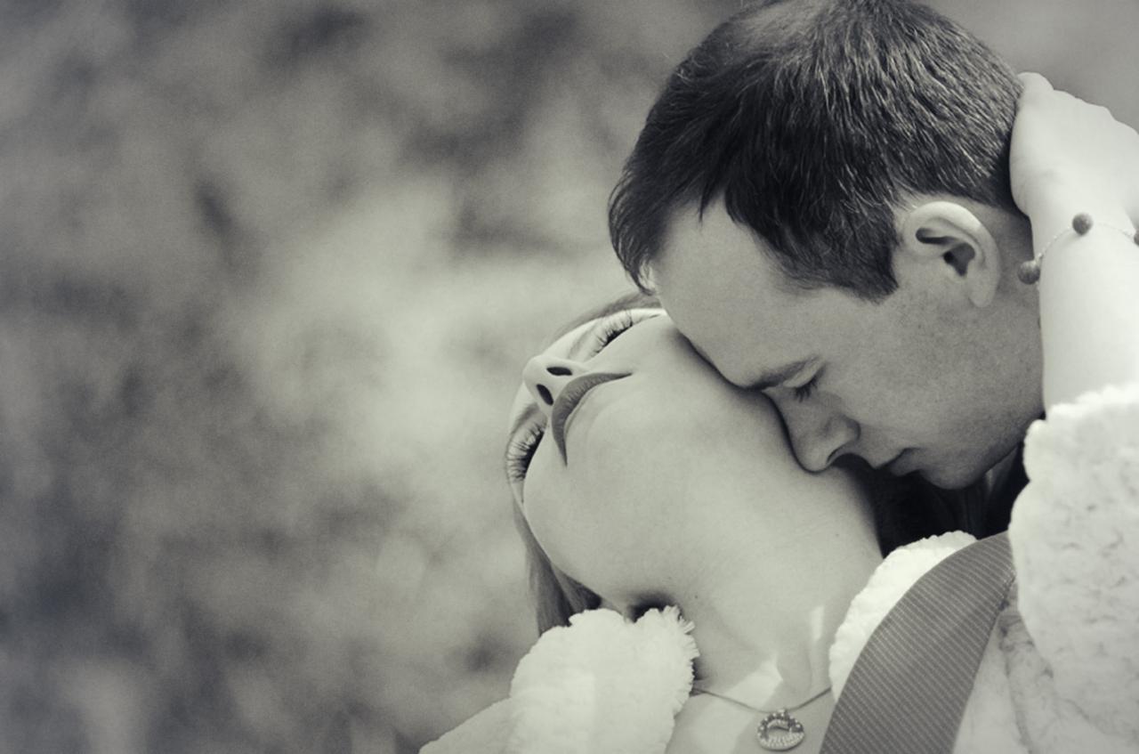 Czarno-białe zbliżenie całującej się pary, kobieta z podniesioną głową i delikatnym naszyjnikiem, mężczyzna z zamkniętymi oczami, rozmyte tło.