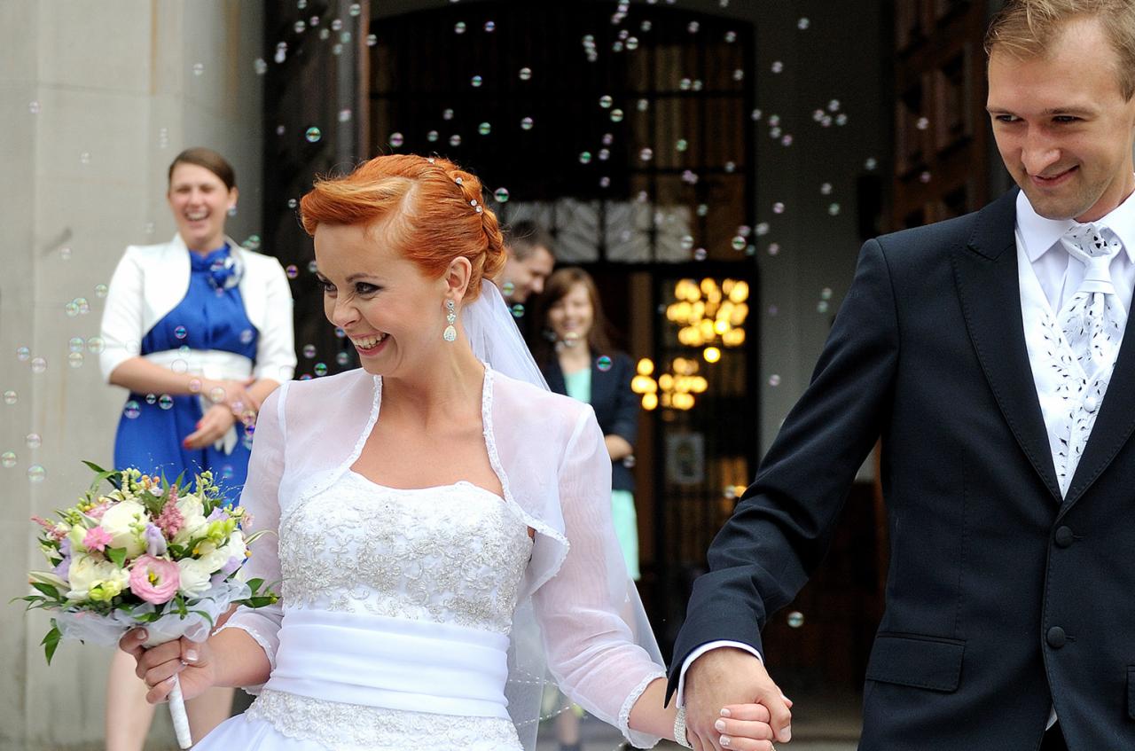 Uśmiechnięta panna młoda z rudymi włosami i bukietem wychodzi z kościoła, trzymając za rękę pana młodego, otoczona bańkami mydlanymi.