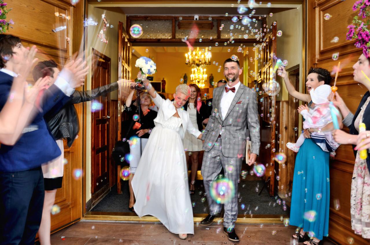 Nowożeńcy, kobieta z bukietem i mężczyzna w szarej kracie, wychodzą z budynku w deszczu baniek mydlanych, otoczeni przez gości.