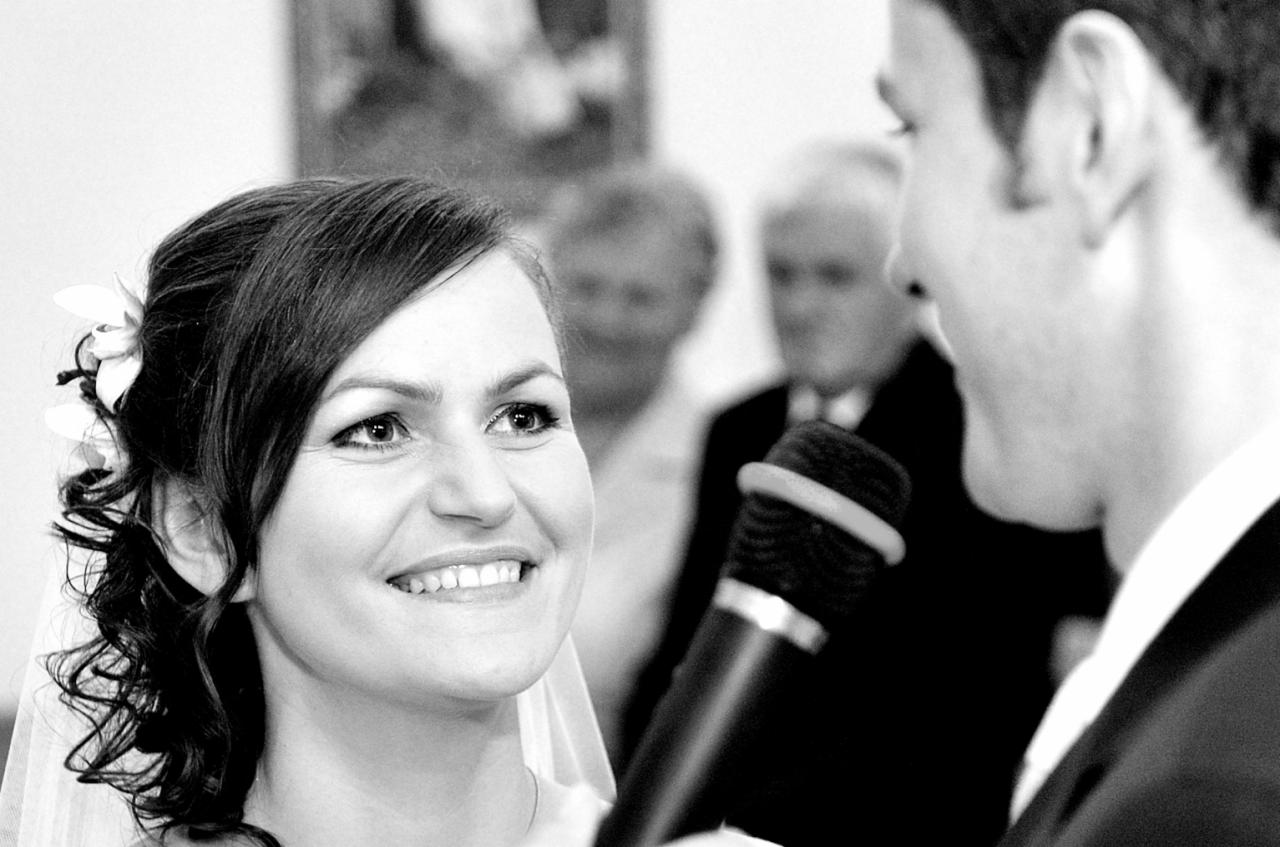 Czarno-białe zbliżenie uśmiechniętej panny młodej z kwiatem we włosach, w tle mężczyzna przy mikrofonie i rozmyte twarze gości.