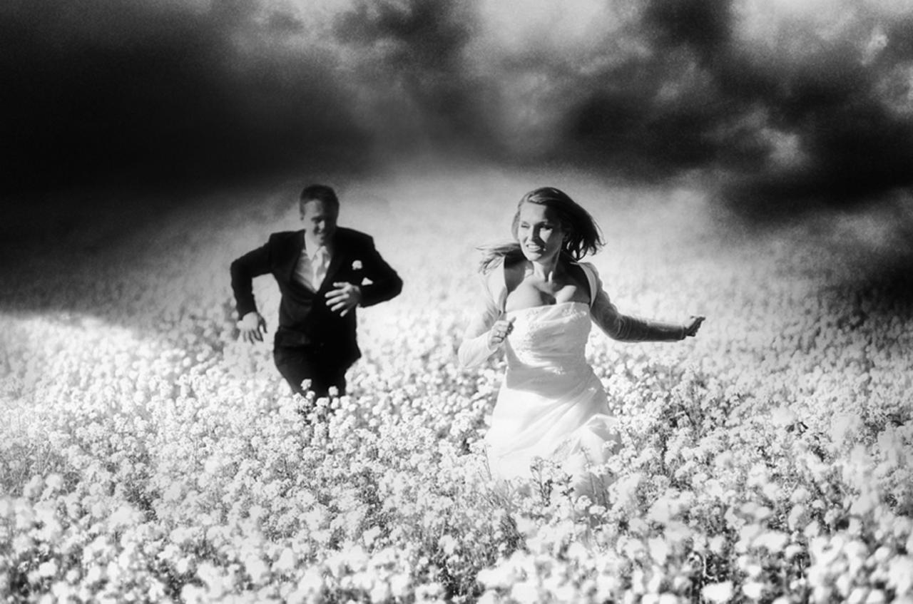 Czarno-białe zdjęcie panny młodej w sukni i pana młodego w garniturze biegnących przez pole kwitnących roślin, z dramatycznym, ciemnym niebem w tle.
