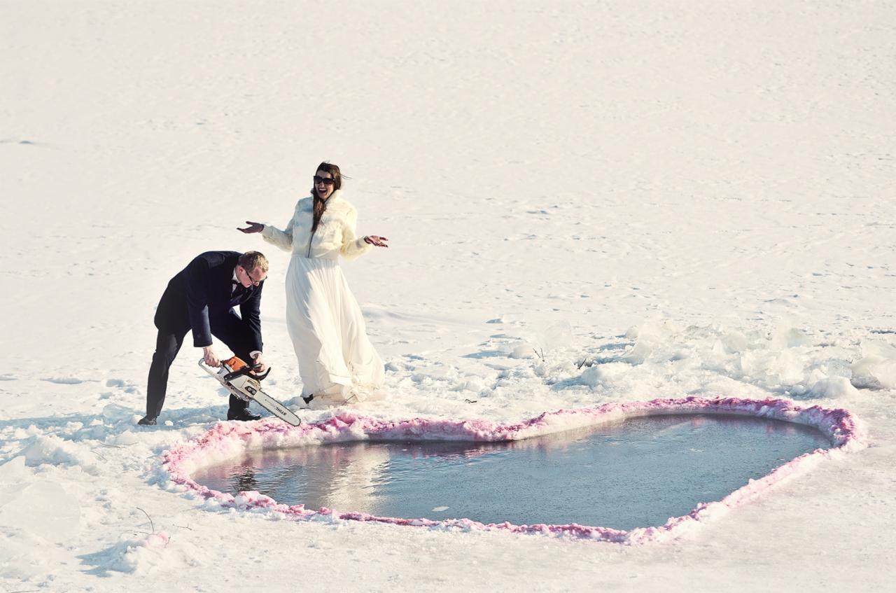 Nietypowa sesja ślubna: Pan młody wycina piłą mechaniczną serce w lodzie, panna młoda w futerku i okularach przeciwsłonecznych stoi obok z rozłożonymi rękami na tle białego śniegu.