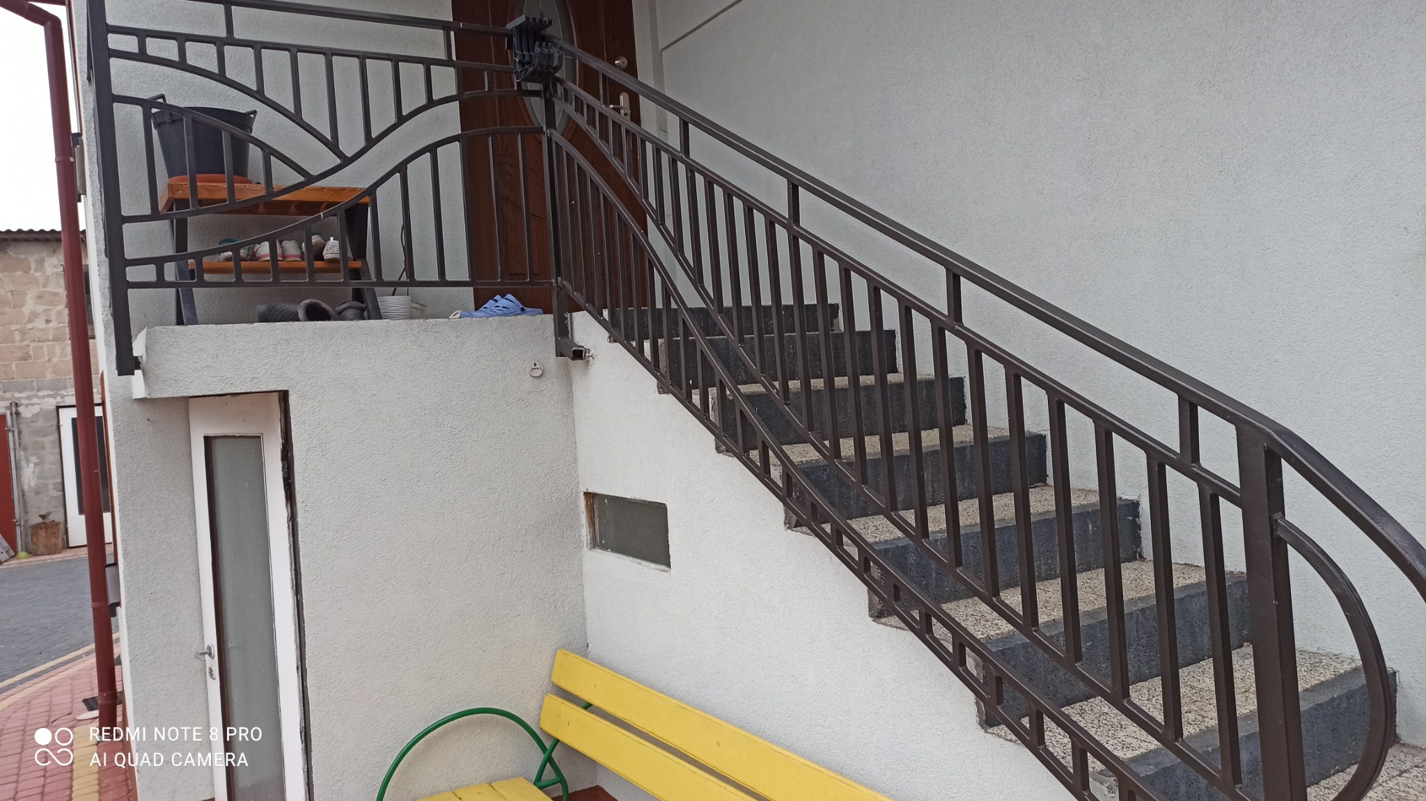 Zewnętrzne schody z metalową balustradą w kolorze ciemnego brązu, prowadzące do drzwi wejściowych. Na górze schodów widoczny balkon z metalową barierką i półką z przedmiotami.