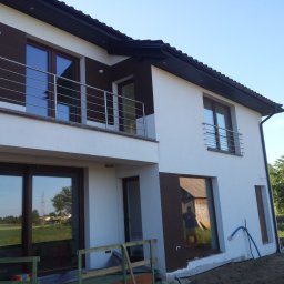 Kuźnia Macieja - Nowoczesny dom jednorodzinny z balkonem zabezpieczonym prostą, poziomą balustradą ze stali nierdzewnej, widoczne okna z ciemnymi ramami i fragment ogrodu.