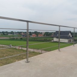 Kuźnia Macieja - Nowoczesna, minimalistyczna balustrada ze stali nierdzewnej z poziomymi prętami, zamontowana na betonowym tarasie z widokiem na wiejski krajobraz z domami w tle.