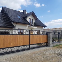 Kuźnia Macieja - Nowoczesna brama przesuwna z drewnianym wypełnieniem i ozdobnymi elementami kutymi, z widocznym fragmentem domu z czarnym dachem i budową w tle.