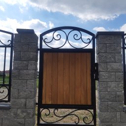 Kuźnia Macieja - Brama wjazdowa z drewnianym wypełnieniem i kutymi elementami, osadzona na słupkach z szarej kostki, na tle błękitnego nieba z chmurami.
