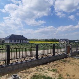 Kuźnia Macieja - Czarny, metalowy płot z ozdobnymi elementami, zbudowany na niskiej, szarej podmurówce z betonowych bloczków, na tle błękitnego nieba z białymi chmurami i żółtego pola rzepaku w oddali.