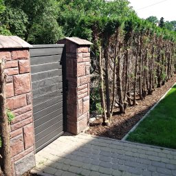 Kuźnia Macieja - Nowoczesna brama wjazdowa z poziomych paneli, osadzona na murowanych słupkach z okładziny imitującej kamień, otoczona żywopłotem i brukowanym podjazdem.