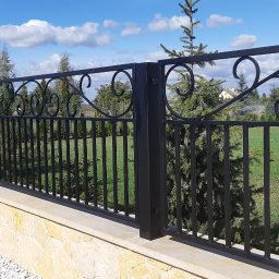 Kuźnia Macieja - Czarna, metalowa konstrukcja ogrodzeniowa z dekoracyjnymi elementami w kształcie spirali, zamontowana na niskim murku z kamienia naturalnego w Świdniku. W tle widoczny zadbany ogród i błękitne...