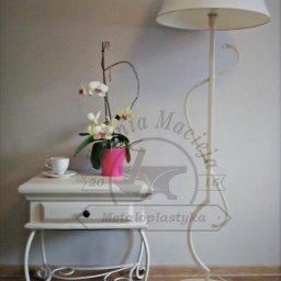 Kuźnia Macieja - Stylowa, metalowa lampa podłogowa w kolorze ecru z dekoracyjnym, spiralnym elementem, stojąca obok białego, metalowego stolika z szufladą, na którym znajduje się filiżanka i doniczka z orchideą.