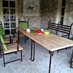 Kuźnia Macieja - Rustykalny stół z drewnianym blatem i metalową podstawą, otoczony krzesłami z zielonymi poduszkami, ustawiony na kamiennym tarasie z widocznym logo firmy 'Kuznia Macieja Metaloplastyka' na ścianie...