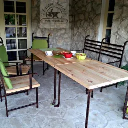 Rustykalny stół z drewnianym blatem i metalową podstawą, otoczony krzesłami z zielonymi poduszkami, ustawiony na kamiennym tarasie z widocznym logo firmy 'Kuznia Macieja Metaloplastyka' na ścianie...