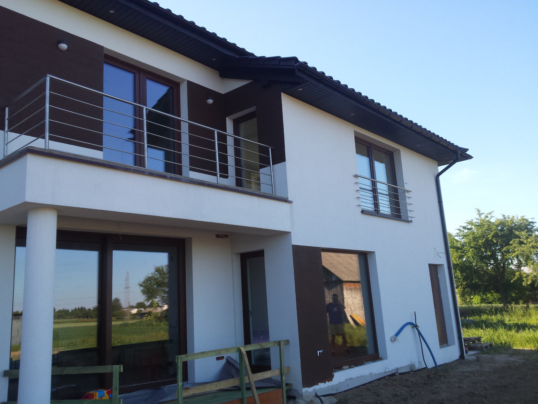 Nowoczesny dom jednorodzinny z balkonem zabezpieczonym prostą, poziomą balustradą ze stali nierdzewnej, widoczne okna z ciemnymi ramami i fragment ogrodu.