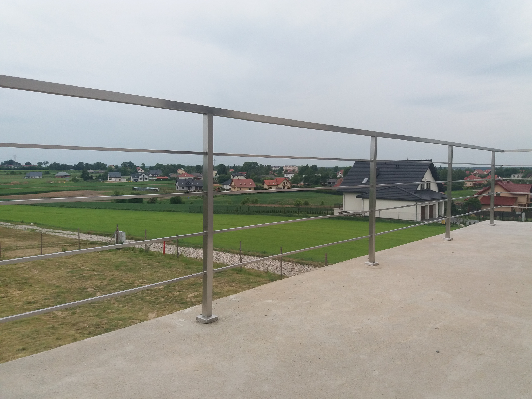 Nowoczesna, minimalistyczna balustrada ze stali nierdzewnej z poziomymi prętami, zamontowana na betonowym tarasie z widokiem na wiejski krajobraz z domami w tle.