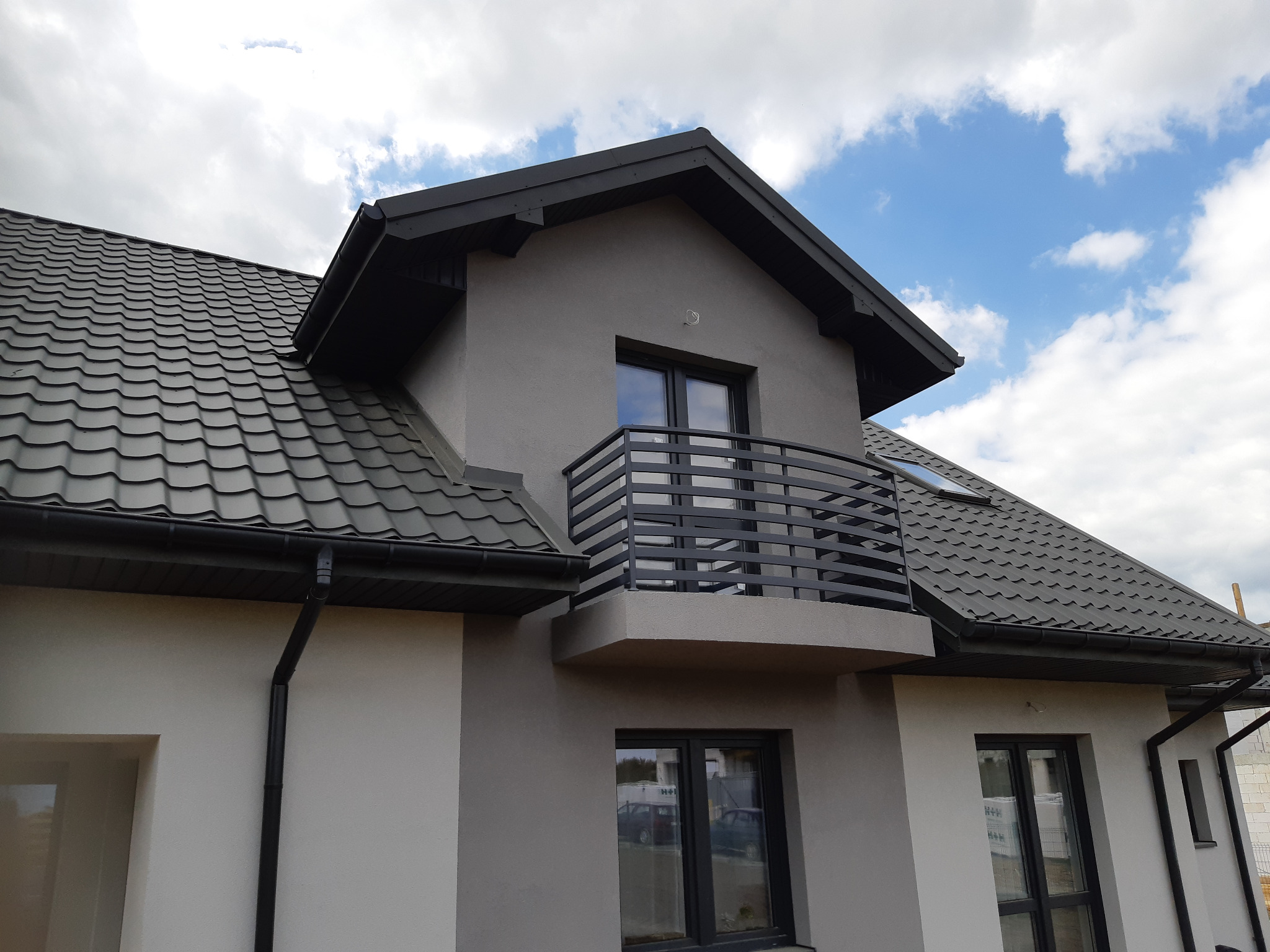 Szary dom jednorodzinny z czarną, poziomą balustradą balkonową, ciemną dachówką i orynnowaniem, uchwycony w słoneczny dzień z lekkim zachmurzeniem.