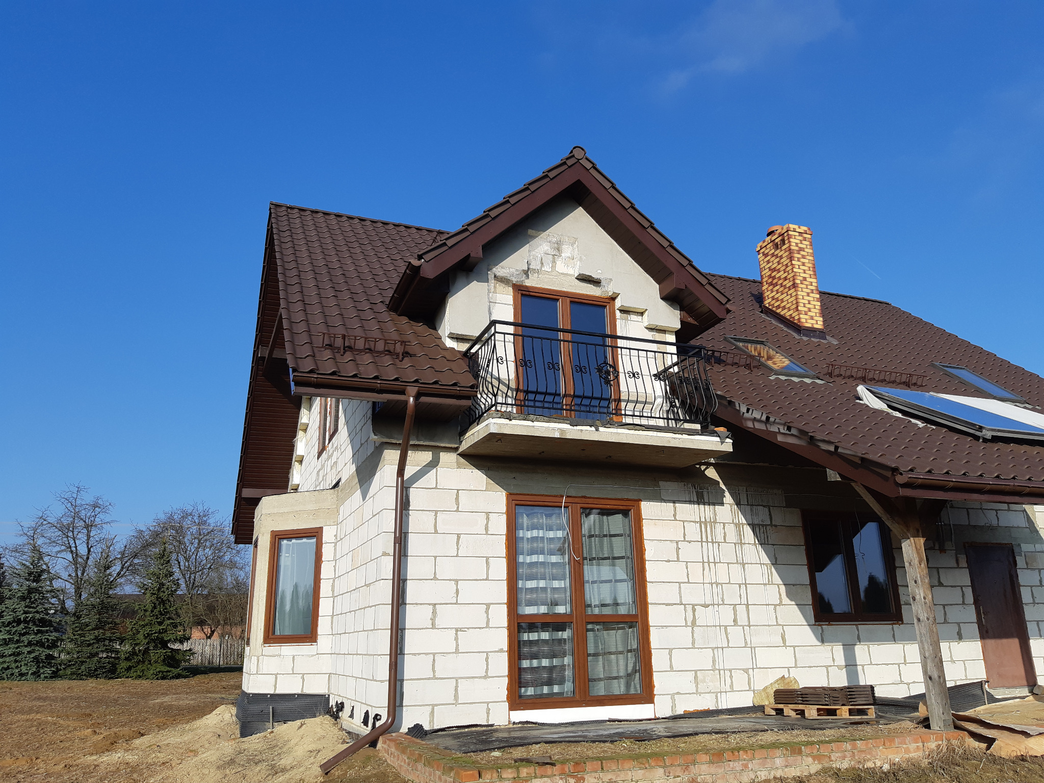 Surowy stan domu jednorodzinnego w trakcie budowy, z widocznym balkonem z czarną, kutą balustradą, brązową dachówką i białymi ścianami z bloczków, na tle błękitnego nieba.