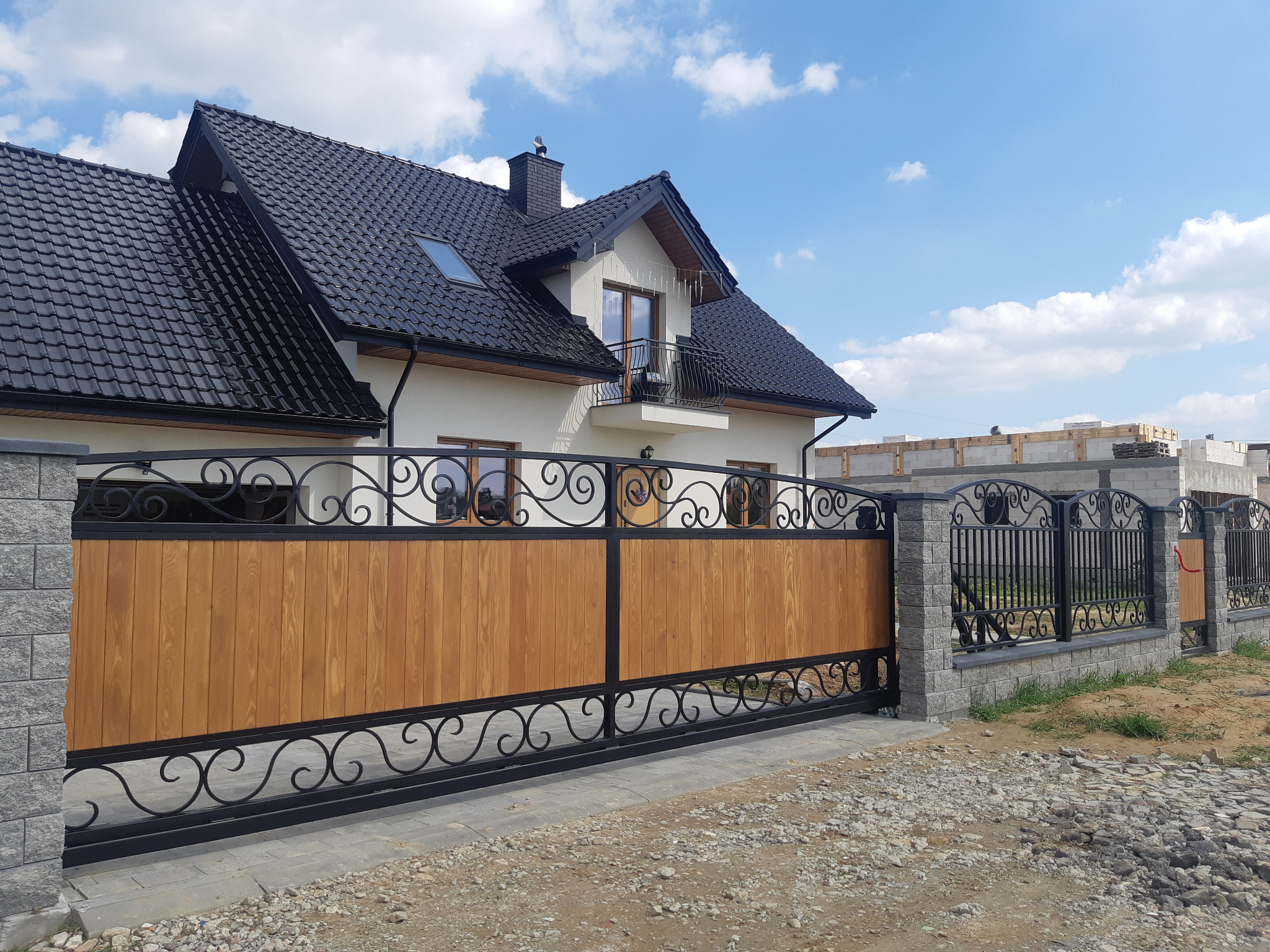 Nowoczesna brama przesuwna z drewnianym wypełnieniem i ozdobnymi elementami kutymi, z widocznym fragmentem domu z czarnym dachem i budową w tle.