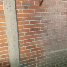 Fragment ceglanej ściany z widocznymi śladami po piaskowaniu, z lewej strony ściana z cegły po prawej po piaskowaniu, ślad po metalowym elemencie na górze ściany.