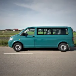 Turkusowy bus Volkswagen Transporter T5 zaparkowany na poboczu drogi, widok z boku, w tle pola i zabudowania.