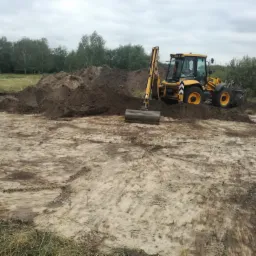Żółta koparko-ładowarka JCB 4CX na placu budowy, z hałdami ziemi w tle i pochmurnym niebem.