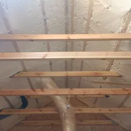 Poddasze ocieplone pianką PUR, widoczne drewniane elementy konstrukcyjne, fragment instalacji wentylacyjnej w folii oraz wisząca żarówka.