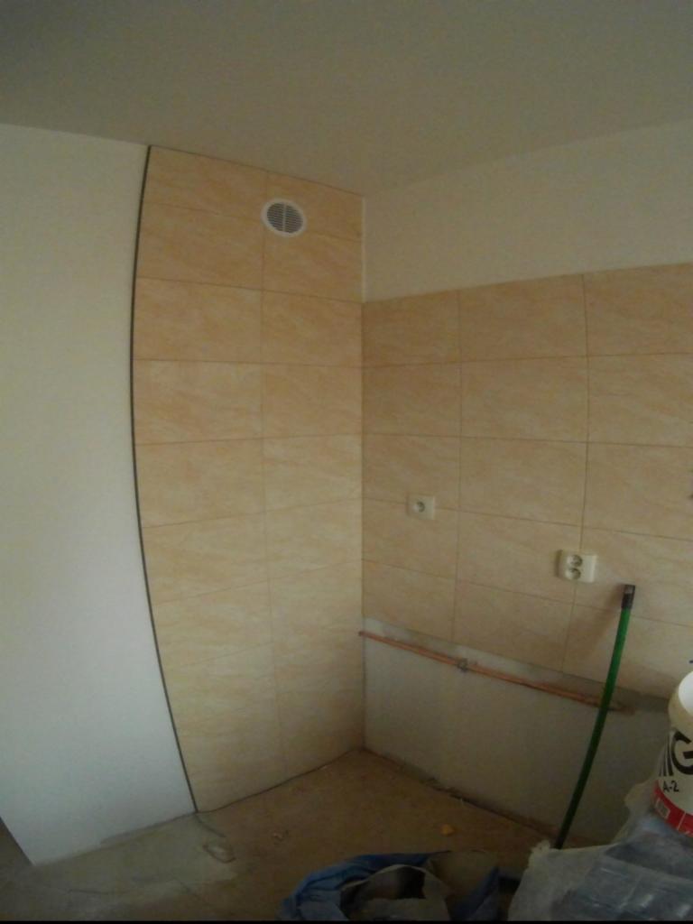 Ściany łazienki w trakcie układania jasnych, beżowych płytek ceramicznych. Widoczny otwór wentylacyjny w górnej części ściany i wystające rury instalacyjne.