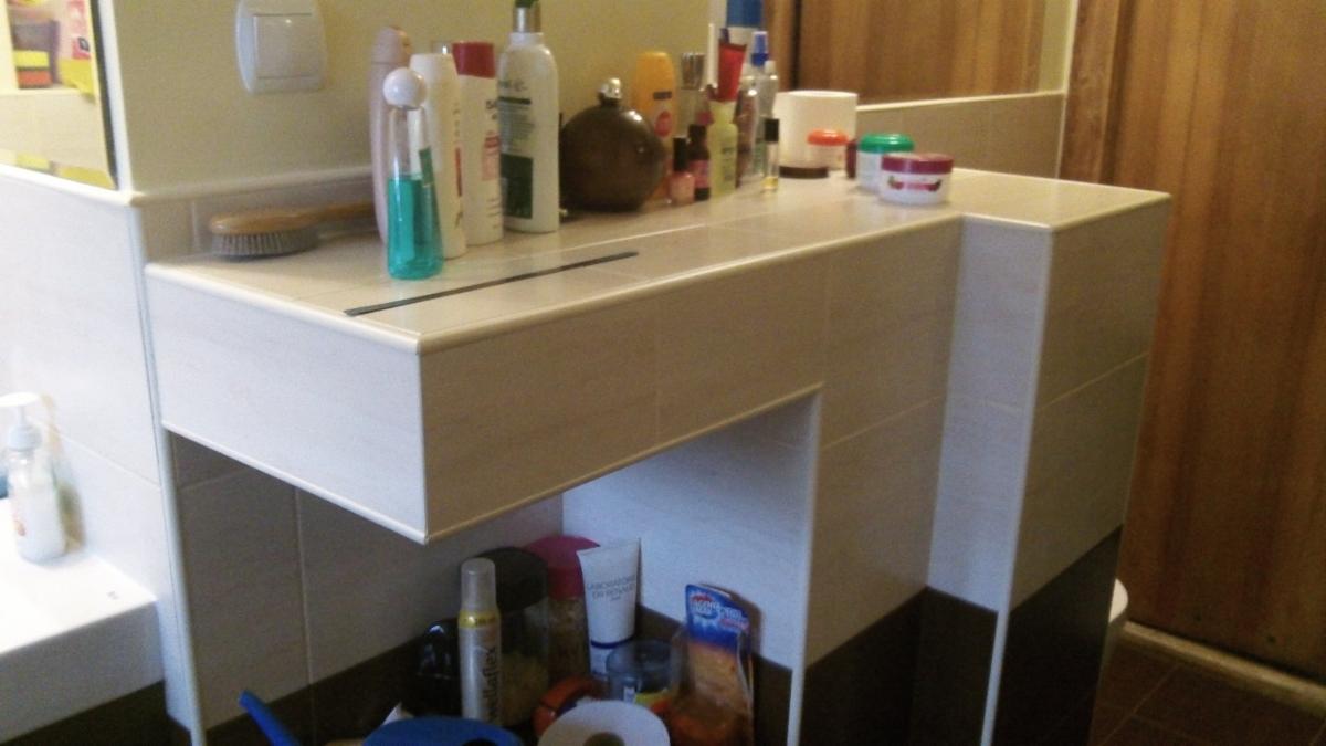 Umywalka z białych płytek z półką pod spodem, na której stoją kosmetyki i środki czystości, na blacie umywalki również ustawione różne butelki i pojemniki.