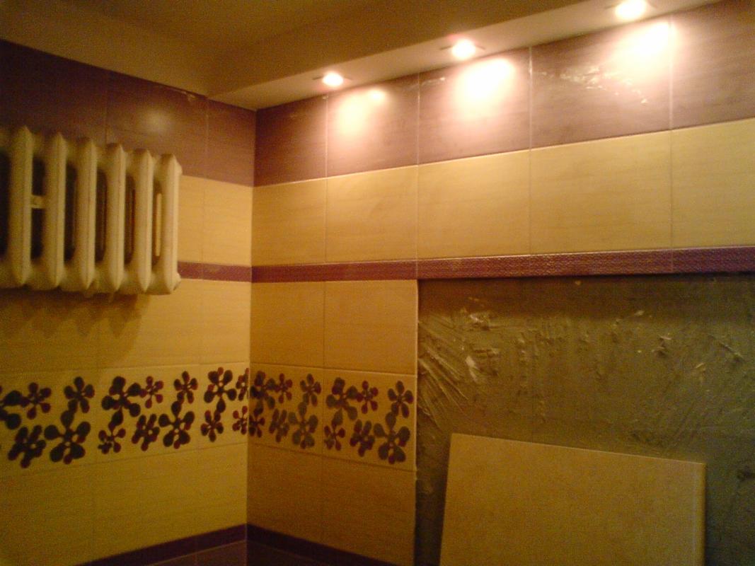 Fragment łazienki z beżowo-brązowymi płytkami, dekoracyjnym pasem z motywem kwiatowym i grzejnikiem żeberkowym; widoczny fragment ściany w trakcie prac wykończeniowych.