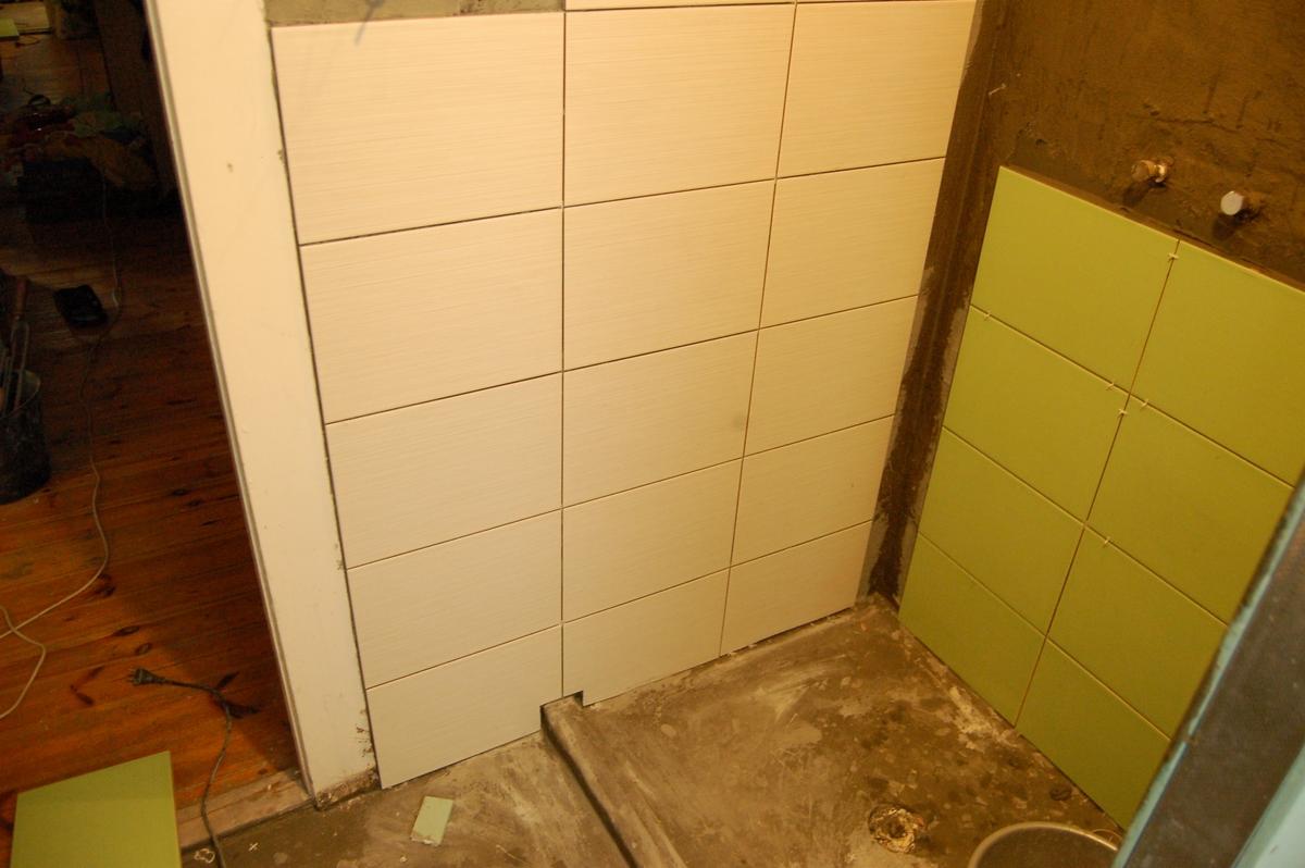 Ściana łazienki w trakcie układania jasnych, podłużnych płytek ceramicznych, kontrastująca z fragmentem ściany wyłożonej zielonymi płytkami w tle, widoczne niedoskonałości podłoża i brak fug.