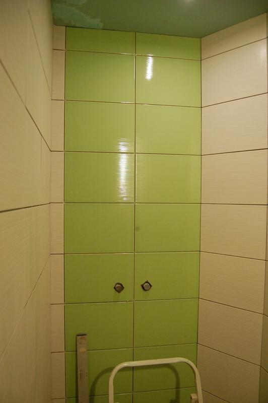 Ściana w łazience wyłożona błyszczącymi, pistacjowymi płytkami ceramicznymi z widocznymi otworami na baterię oraz fragmentem drabinki i poziomicy.