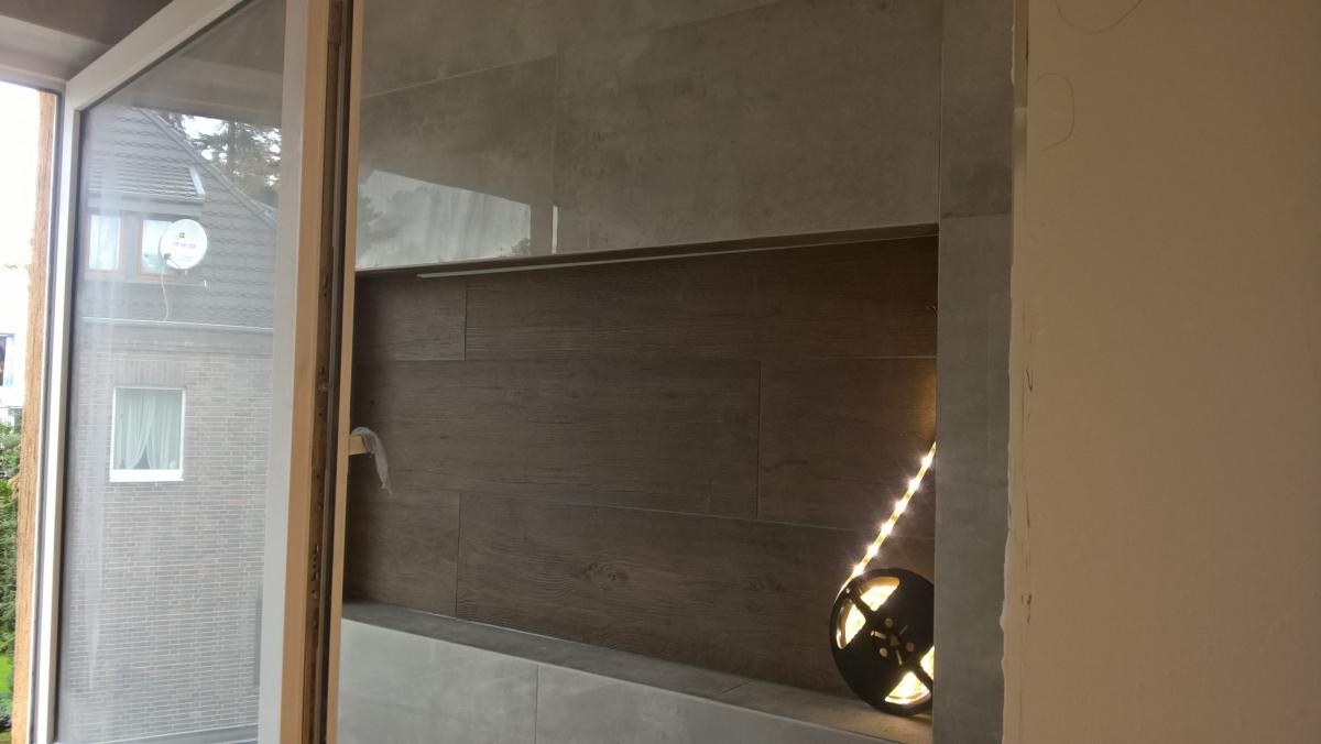 Wykończona wnęka w łazience z szarymi płytkami imitującymi drewno, oświetlona listwą LED i dekoracyjną lampką, widok z okna na budynek mieszkalny.