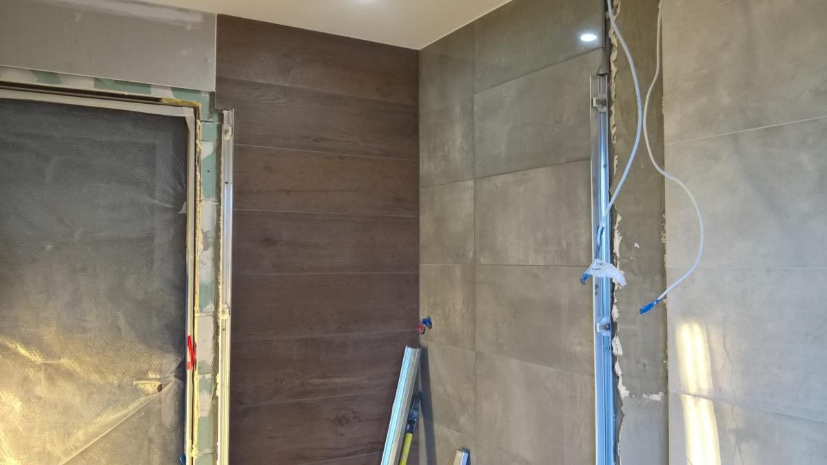 Wnętrze łazienki w trakcie remontu: częściowo wyłożone płytkami imitującymi drewno i szarym gresem, widoczne narzędzia i instalacja wodna.