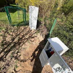Amper-Wolt - Otwarta skrzynka elektryczna z gniazdami na ziemi obok ogrodzenia i krzewów. Widoczne kable i elementy instalacji. Ujęcie z bliska, w tle zieleń i błękit nieba.